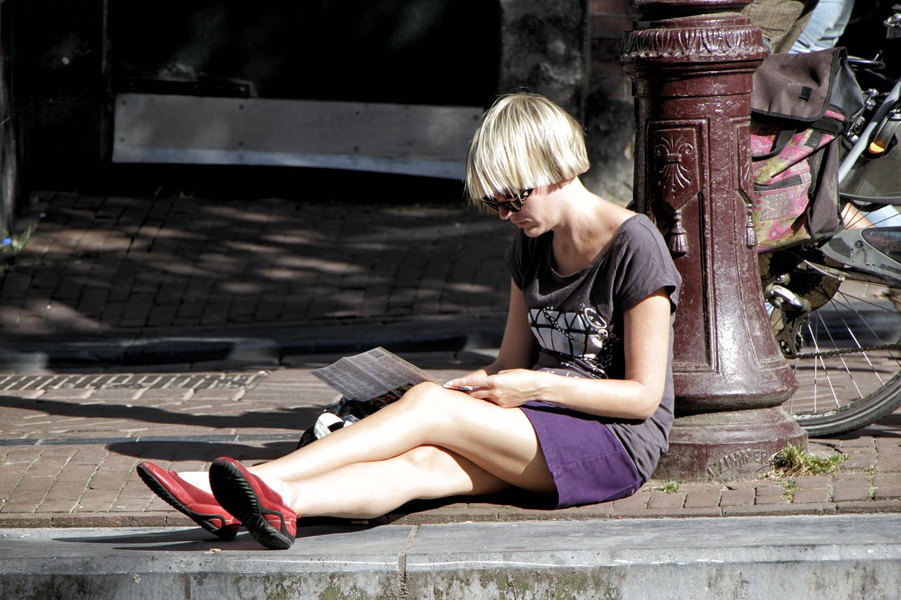 A person with short blonde hair and sunglasses sits on a brick sidewalk, looking at a document.