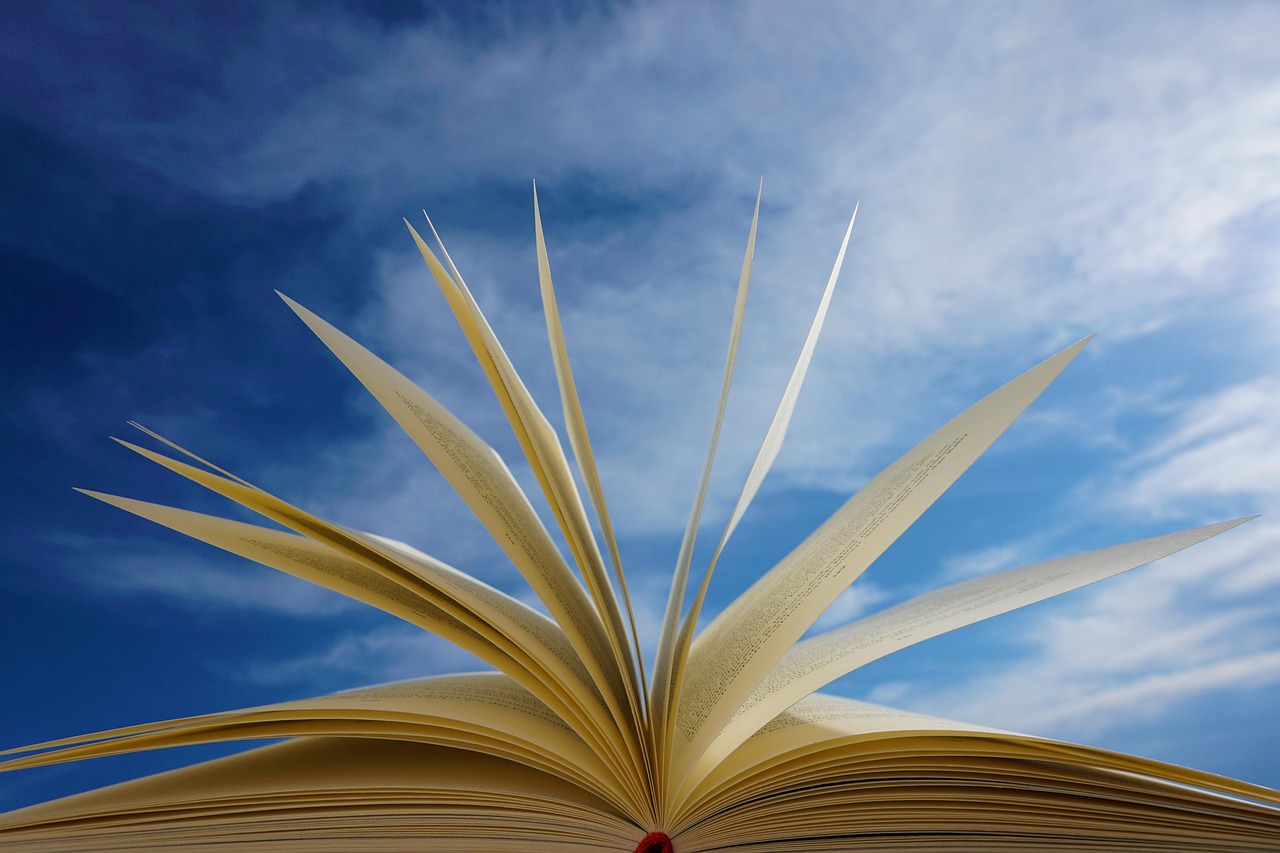An open book with fanned pages against a bright blue sky with scattered clouds.