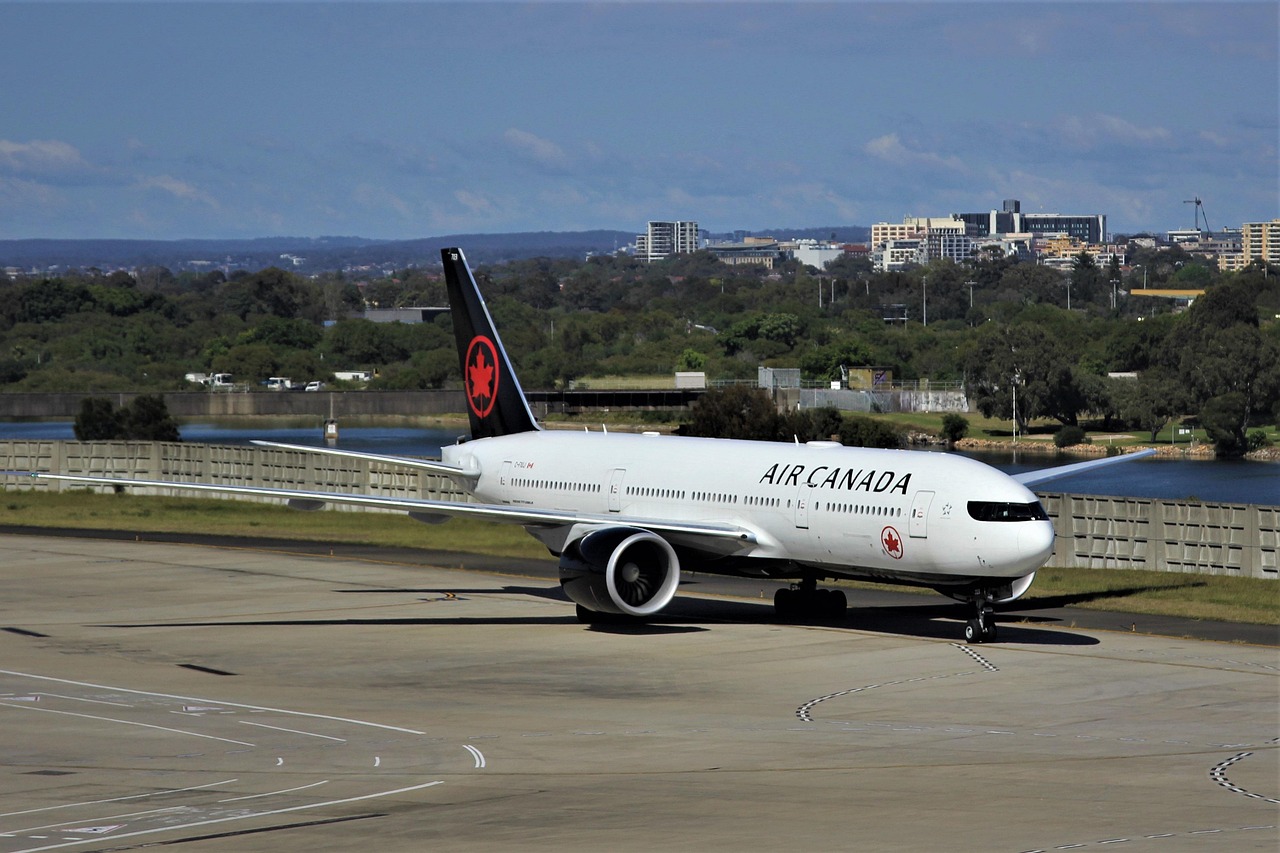A large white Air Canada passenger jet on an airport tarmac with buildings and water in the background.