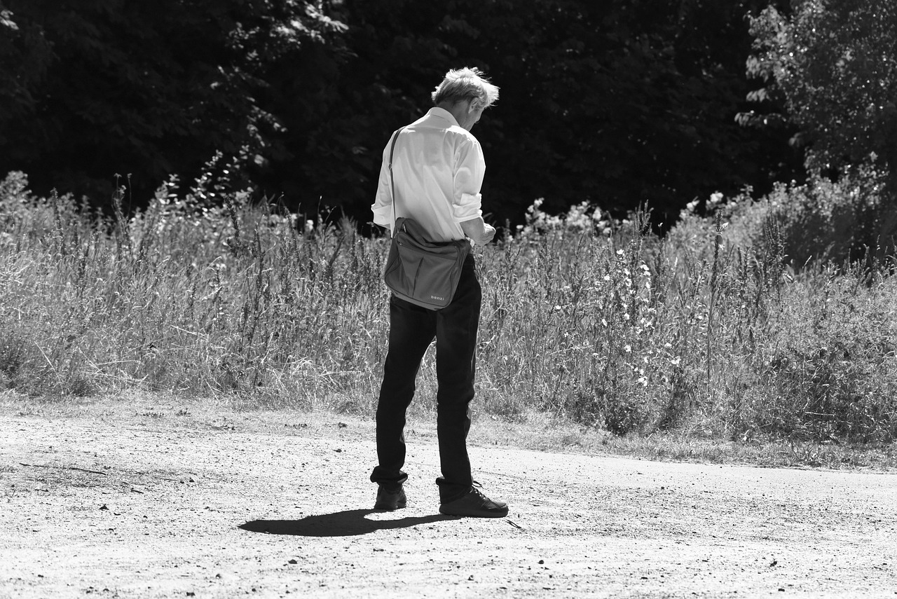 A person in a jacket and pants sits on a wooden park bench, looking down at a small object in their hands.