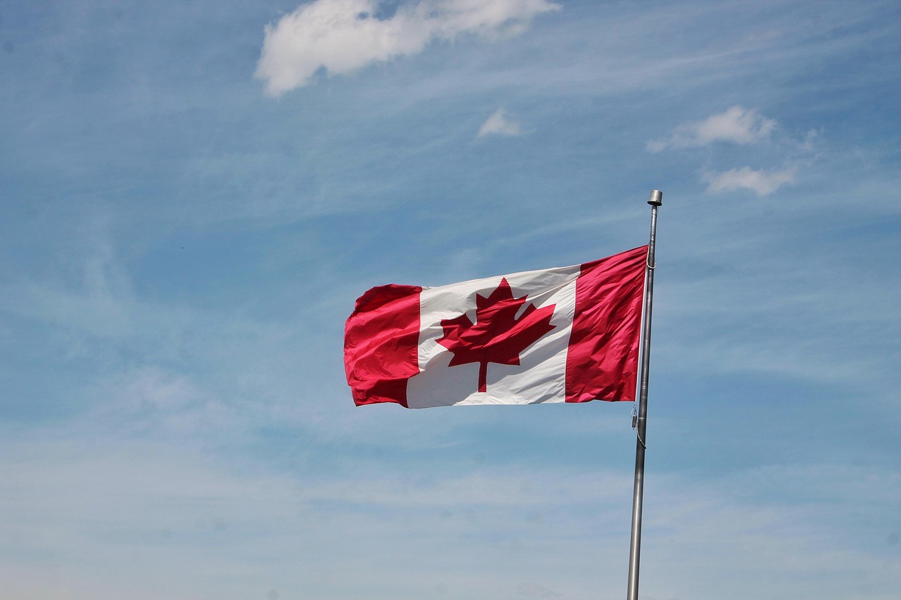 A Canadian flag with a red maple leaf waving on a flagpole against a blue sky with white clouds.