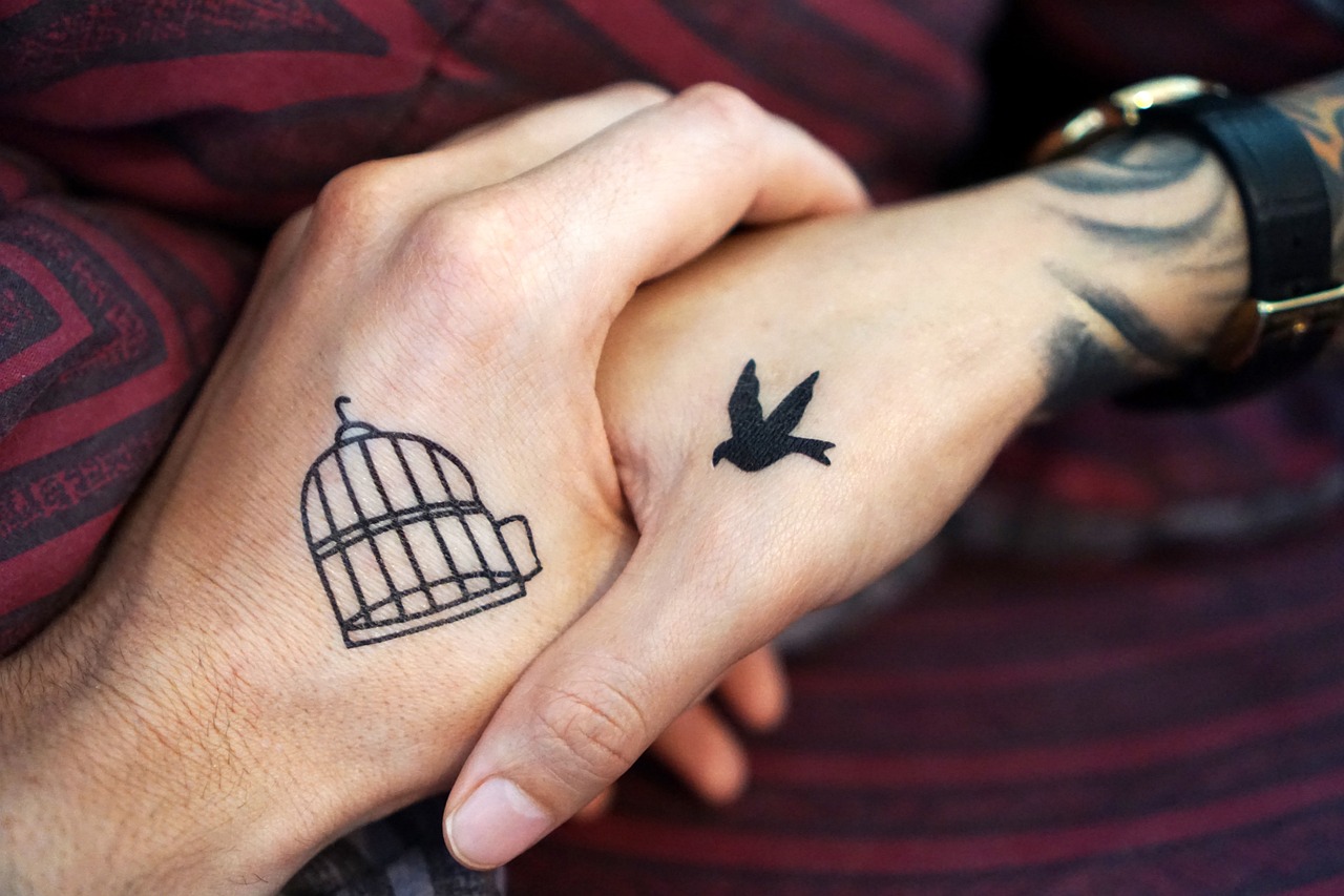 Two hands with a birdcage tattoo and a bird tattoo touching.