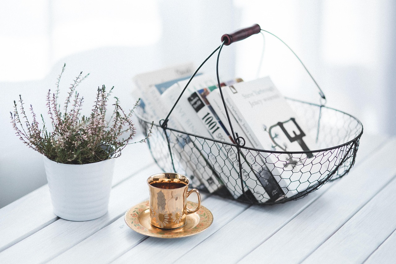 A potted plant, a gold coffee cup, and a wire basket of books on a white wooden table.
