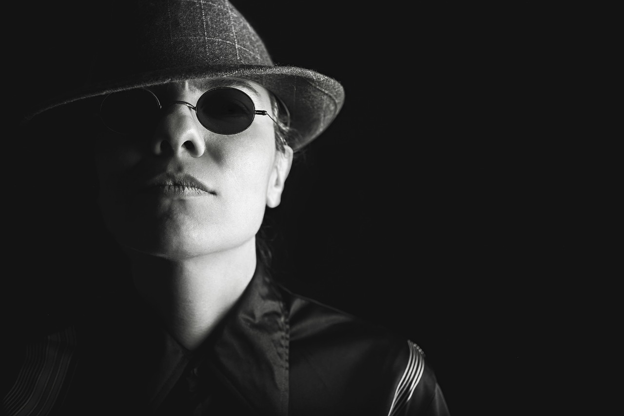 Black and white close-up of a person in a fedora and dark round sunglasses, looking up.