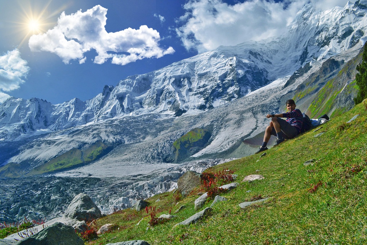 A man sits on a green grassy slope with snow-covered mountains and a glacier in the background under a sunny sky.