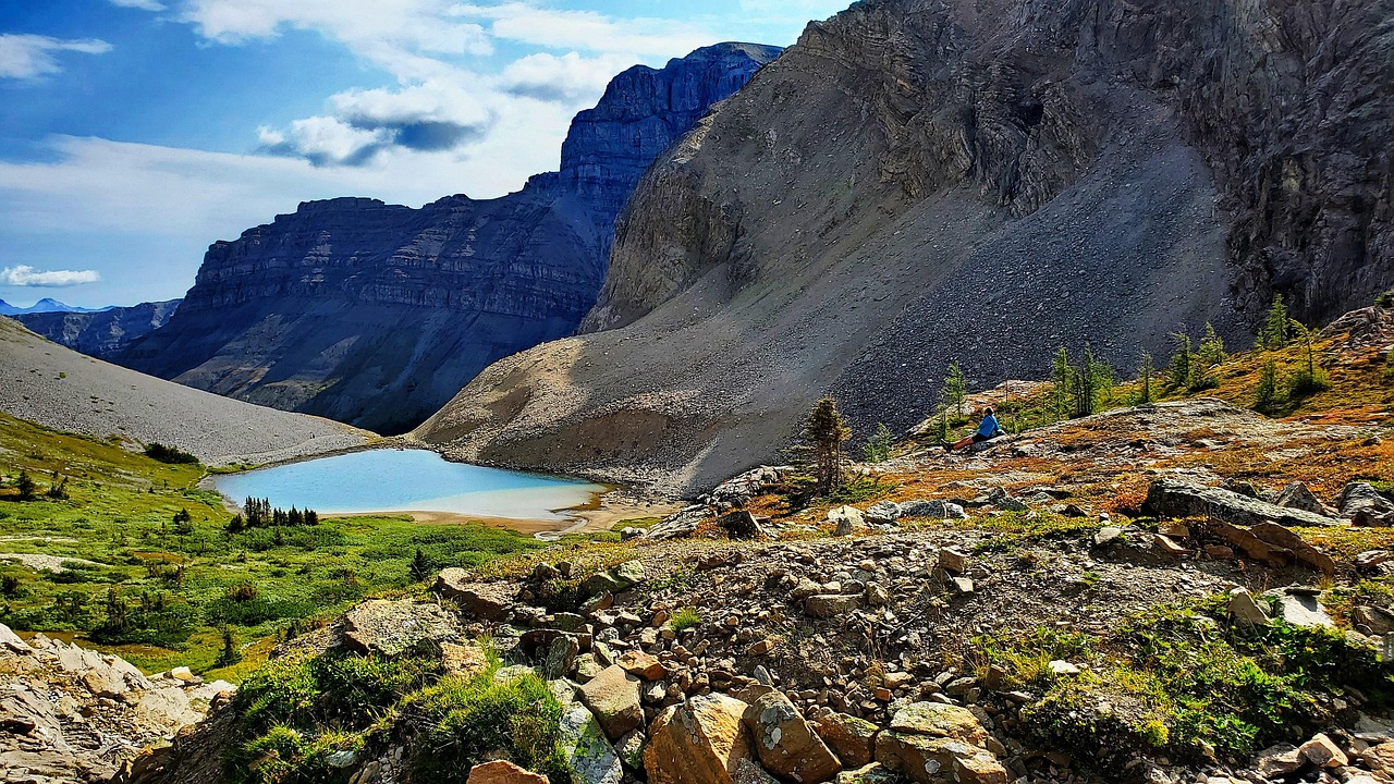A person sits by a small blue lake in a rugged, rocky mountain landscape under a partly cloudy sky.