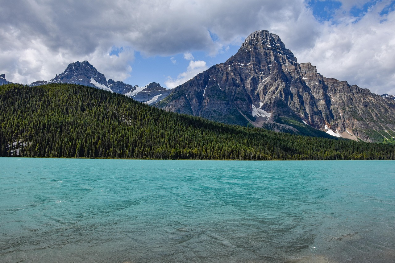 A wide turquoise lake stretches towards a dense evergreen forest at the base of large, snow-capped rocky mountains under a partly cloudy sky.