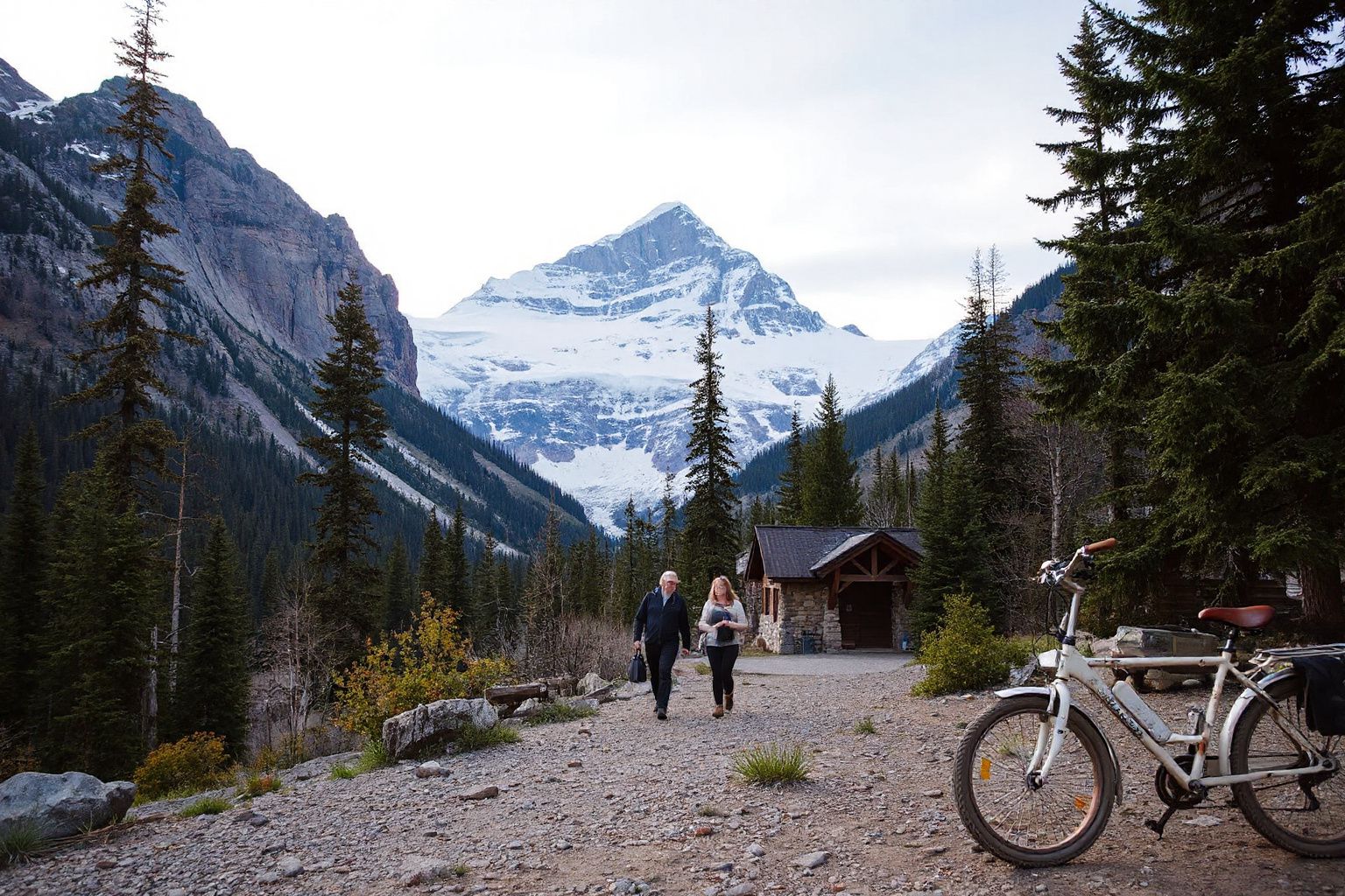 Two people walk on a gravel path towards a stone cabin, with a snow-capped mountain and evergreen forest in the background. A bicycle is parked nearby.