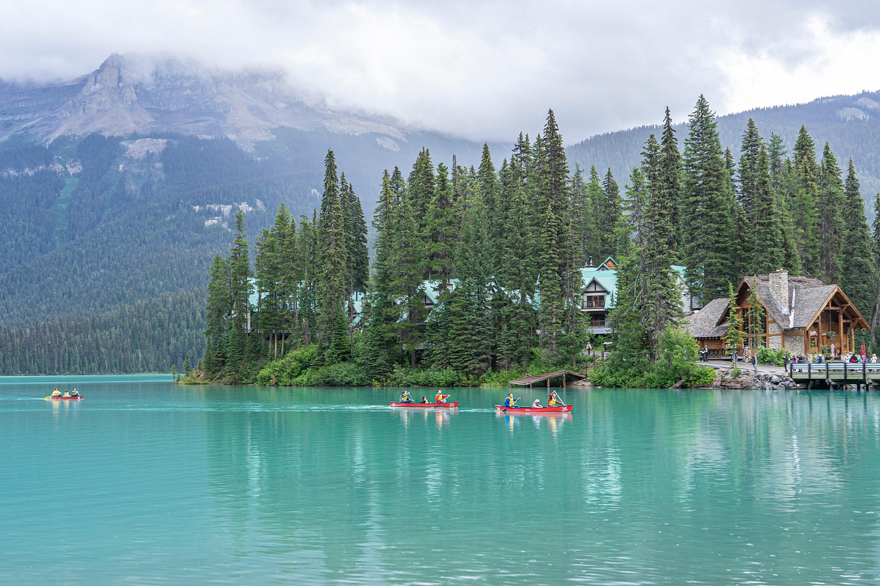 Turquoise lake with canoes, evergreen forest, and rustic lodge buildings under an overcast sky.